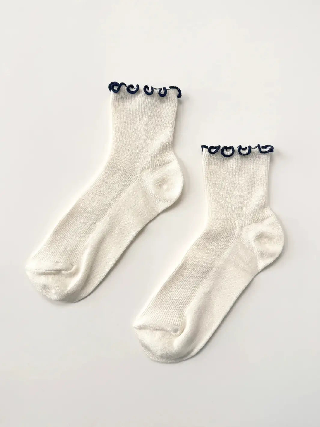 A flat-lay view captures a pair of white and navy Petite Ruffled Modal Socks in Soft White / Navy from Le Bon Shoppe resting on a white background | Twentyseven Toronto