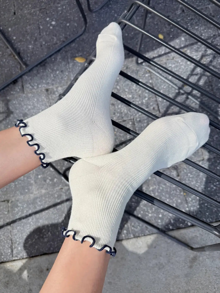 A side view highlights The Petite Ruffled Modal Socks in Soft White / Navy from Le Bon Shoppe being worn on feet resting on a black wire chair | Twentyseven Toronto