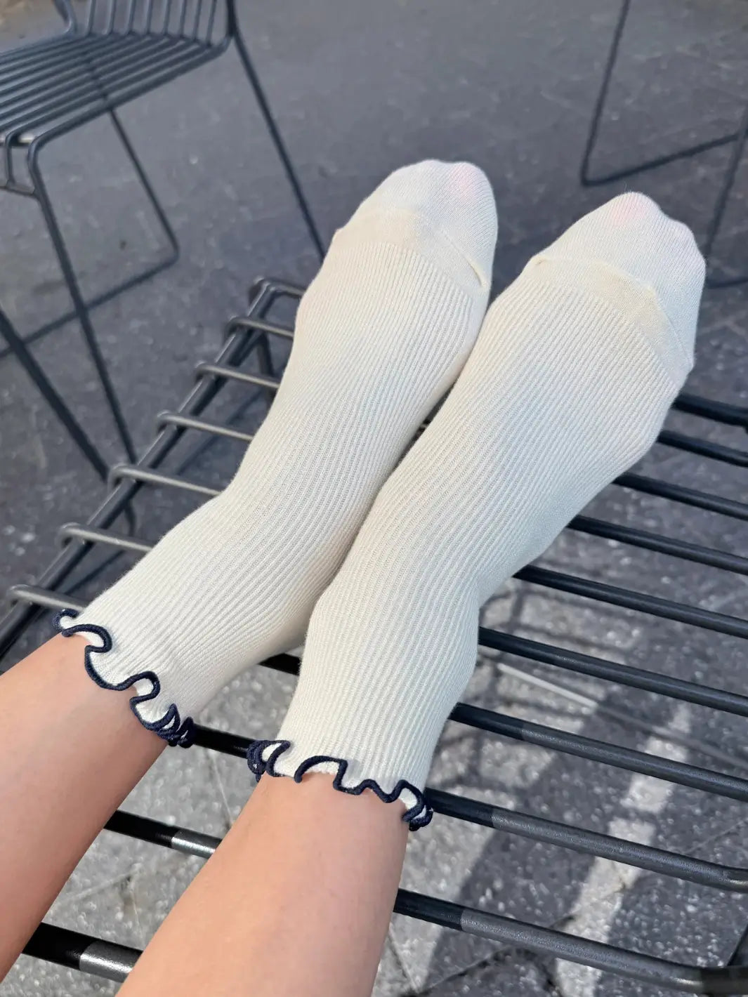 Person wearing white and navy Petite Ruffled Modal Socks in Soft White / Navy from Le Bon Shoppe while resting on a metal chair | Twentyseven Toronto