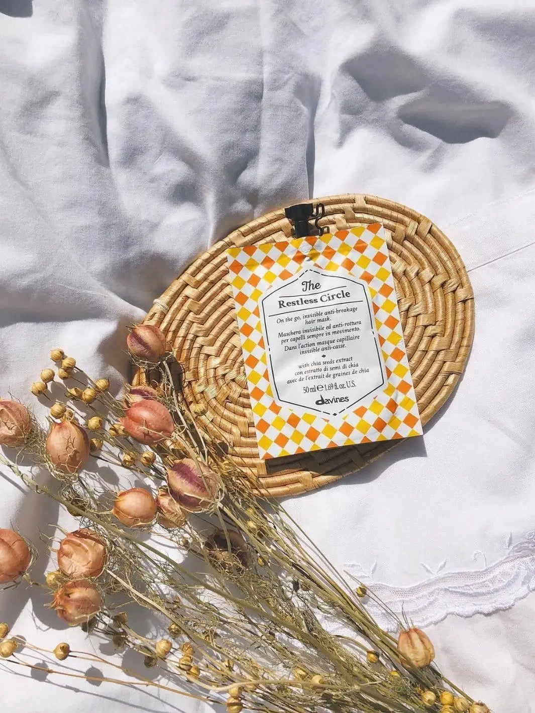 A pouch of Davines The Restless Circle of Chronicles Hair Mask sits on a small woven mat alongside dried floral pods on a white fabric surface | Twentyseven Toronto