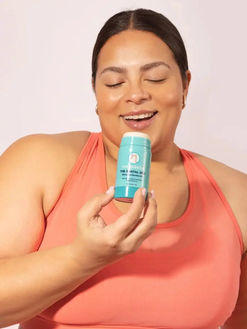 A smiling woman with closed eyes holds a light blue stick of skincare deodorant near her face to highlight the scent of Megababe The Santal Deo | Twentyseven Toornto