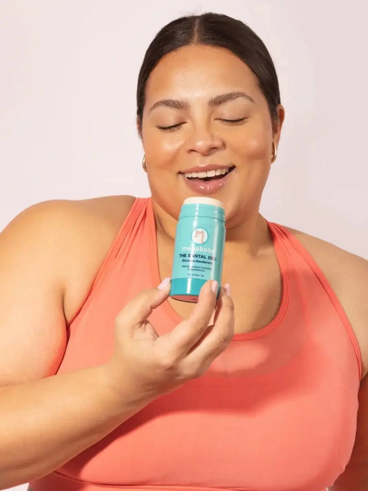 A smiling woman with closed eyes holds a light blue stick of skincare deodorant near her face to highlight the scent of Megababe The Santal Deo | Twentyseven Toornto