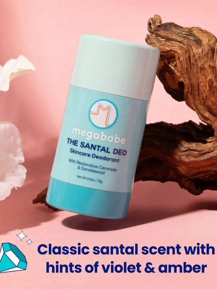 A light blue deodorant container is nestled against a piece of rustic wood on a pink backdrop with text highlighting the scent profile of Megababe The Santal Deo | Twentyseven Toronto
