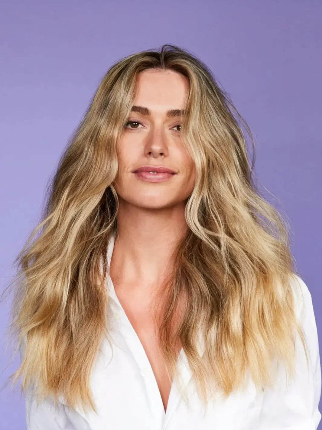 A woman with long, voluminous blonde wavy hair poses against a purple background, showcasing the results achieved using Hair by Sam McKnight The Style Bundle | Twentyseven Toronto