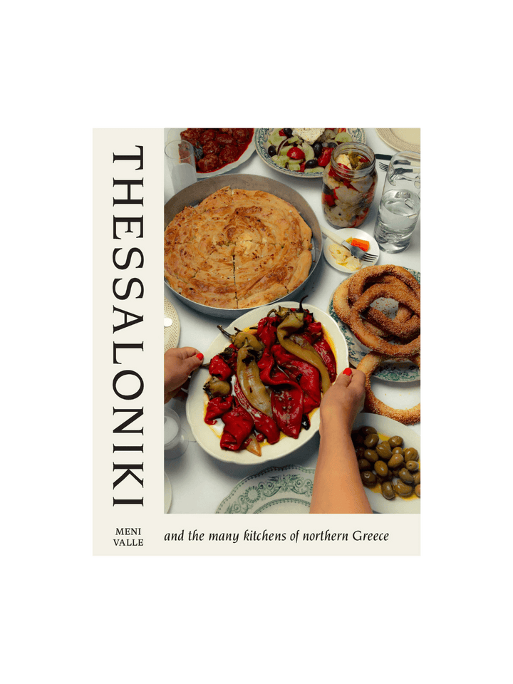 Front cover of the cookbook Thessaloniki: And the Many Kitchens of Northern Greece by Meni Valle, showing a table laden with traditional Greek dishes including roasted red peppers, a large savory pie (pita), olives, and sesame-crusted bread rings (koulouri) | Twentyseven Toronto