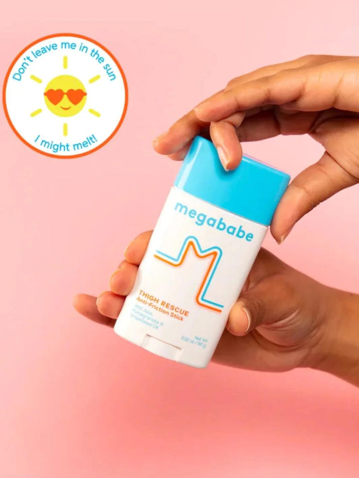 A hand holds up a stick of Megababe Thigh Rescue anti-friction balm against a bright pink background featuring a warning graphic about heat | Twentyseven Toronto