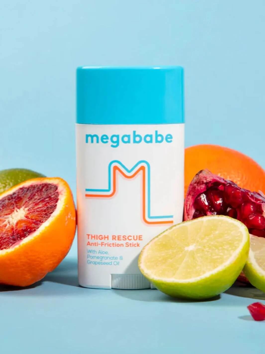 A container of Megababe Thigh Rescue stands on a light blue background surrounded by fresh citrus fruits like limes, blood oranges, and a pomegranate to highlight its natural ingredients | Twentyseven Toronto