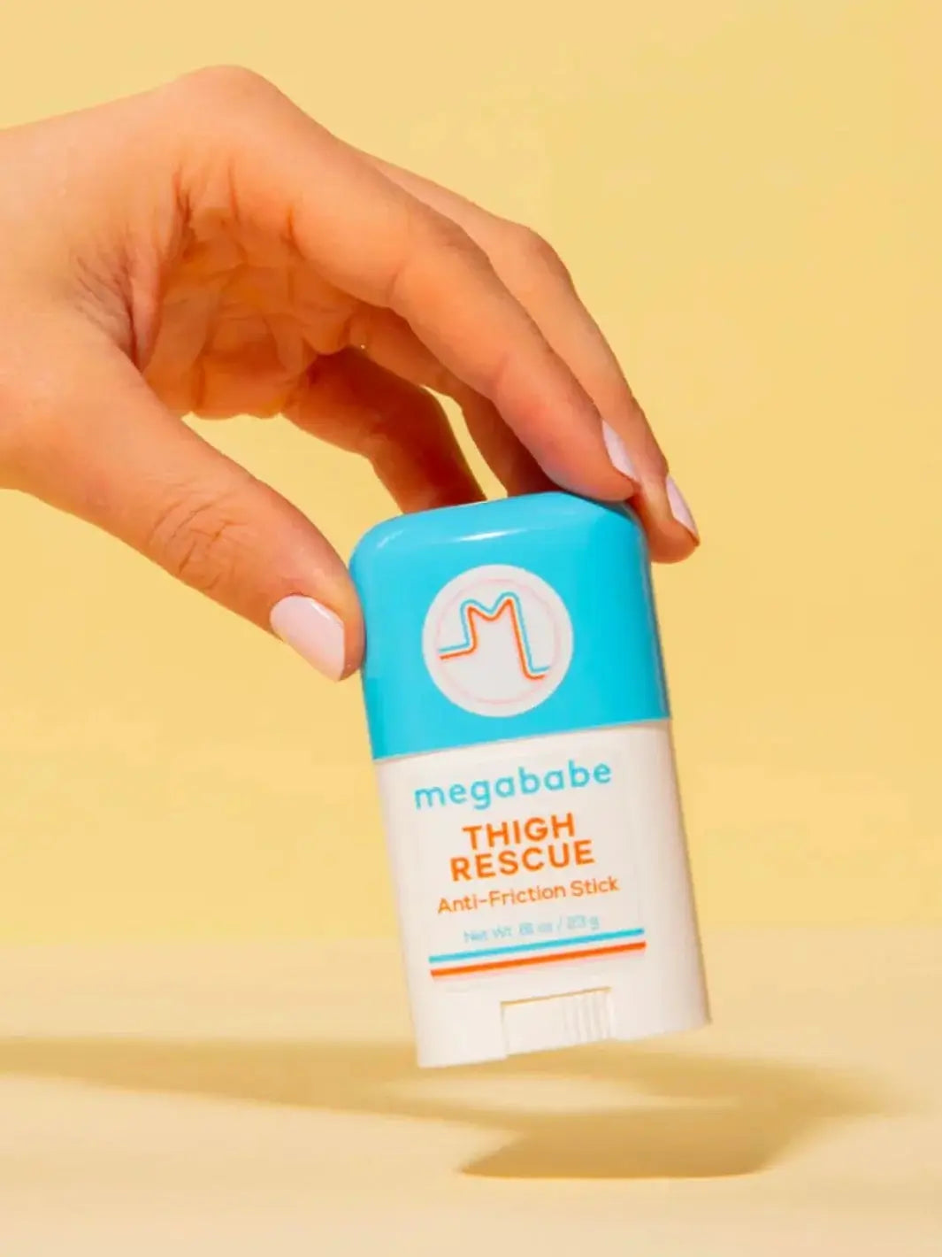 A hand holds the blue and white stick package of Megababe Thigh Rescue Mini against a plain pastel yellow background to emphasize its portable size | Twentyseven Toronto