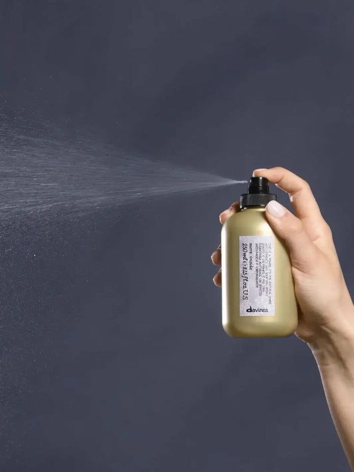 A hand holds a gold bottle of Davines This is a Blow Dry Primer, releasing a fine mist against a solid grey background | Twentyseven Toronto