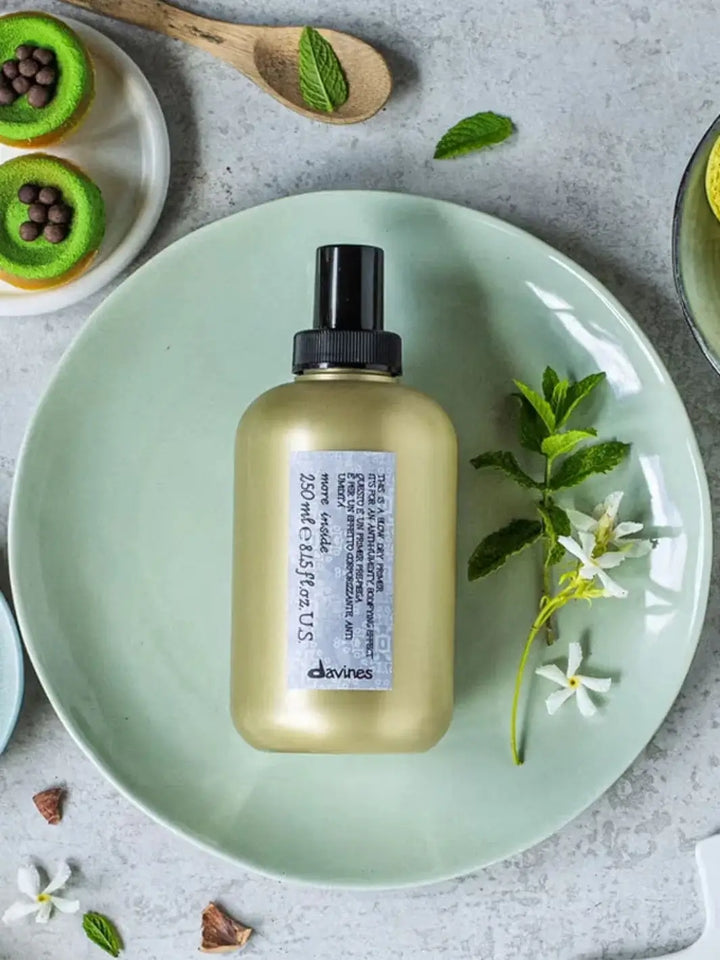 A gold bottle of Davines This is a Blow Dry Primer sits flat on a light green ceramic plate alongside fresh mint leaves and small white flowers | Twentyseven Toronto