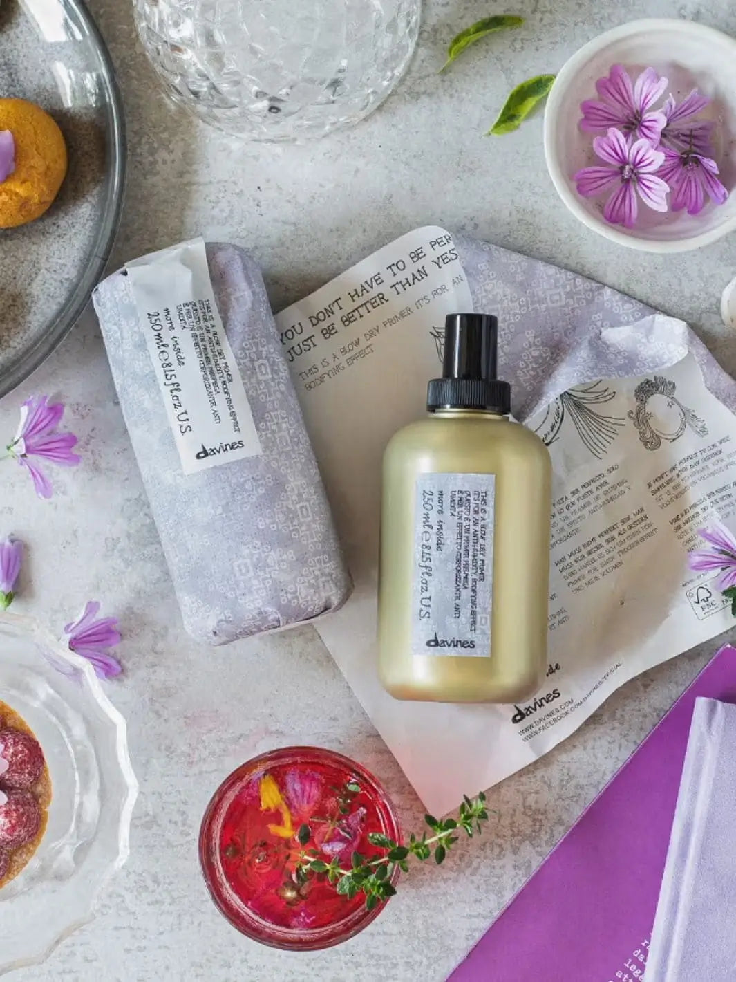 A flat lay arrangement features a gold bottle of Davines This is a Blow Dry Primer next to its patterned purple paper wrapping, surrounded by purple floral accents and decorative treats | Twentyseven Toronto