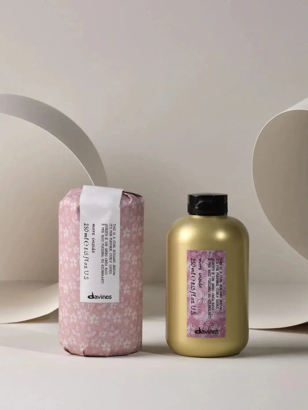 A gold bottle of Davines This is a Curl Building Serum is displayed next to its original pink floral paper packaging against a beige background | Twentyseven Toronto
