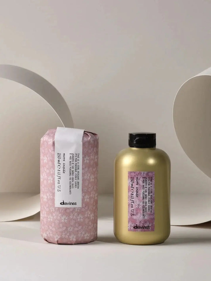 A gold bottle of Davines This is a Curl Building Serum is displayed next to its original pink floral paper packaging against a beige background | Twentyseven Toronto