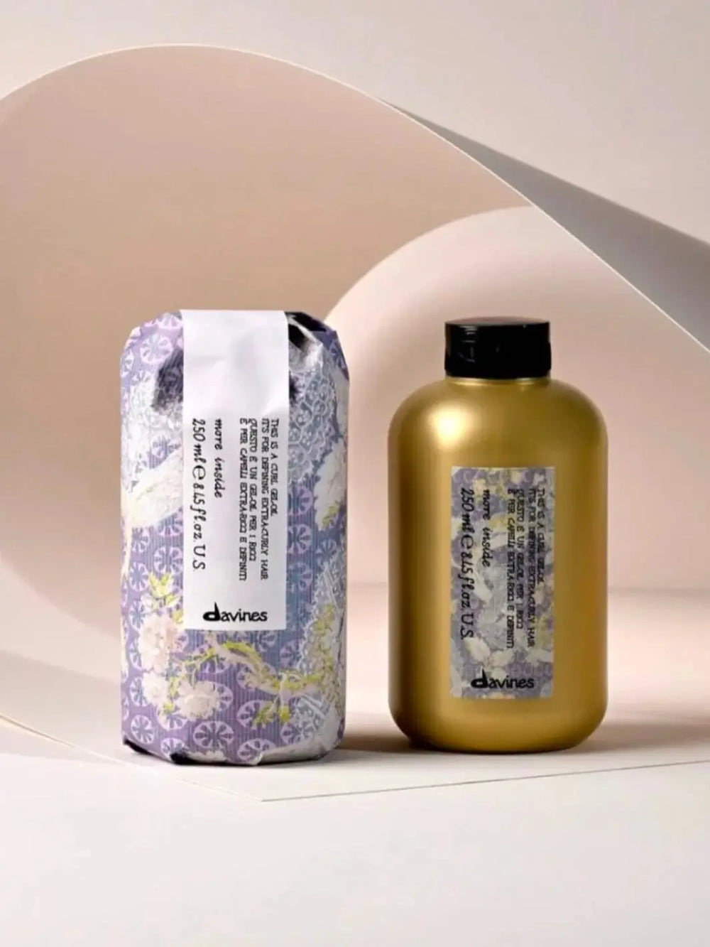 A bottle of Davines This is a Curl Gel Oil stands next to its decorative, purple-patterned paper wrapper against a minimalist beige background | Twentyseven Toronto