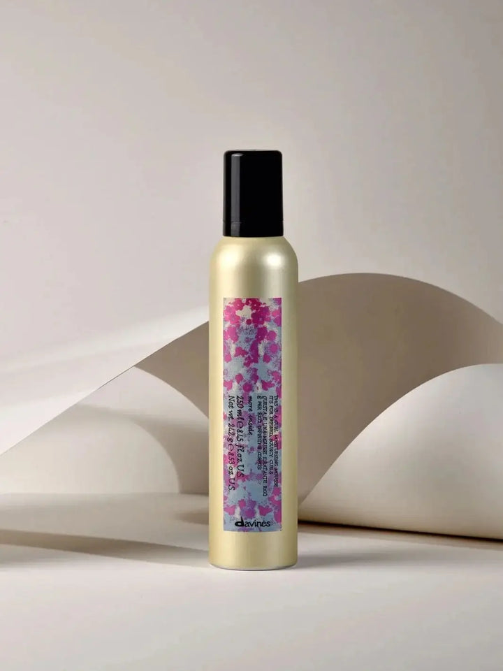 A gold aerosol can of Davines This is a Curl Moisturizing Mousse with a pink and purple abstract floral label stands vertically against a minimalist background of curved white paper | Twentyseven Toronto