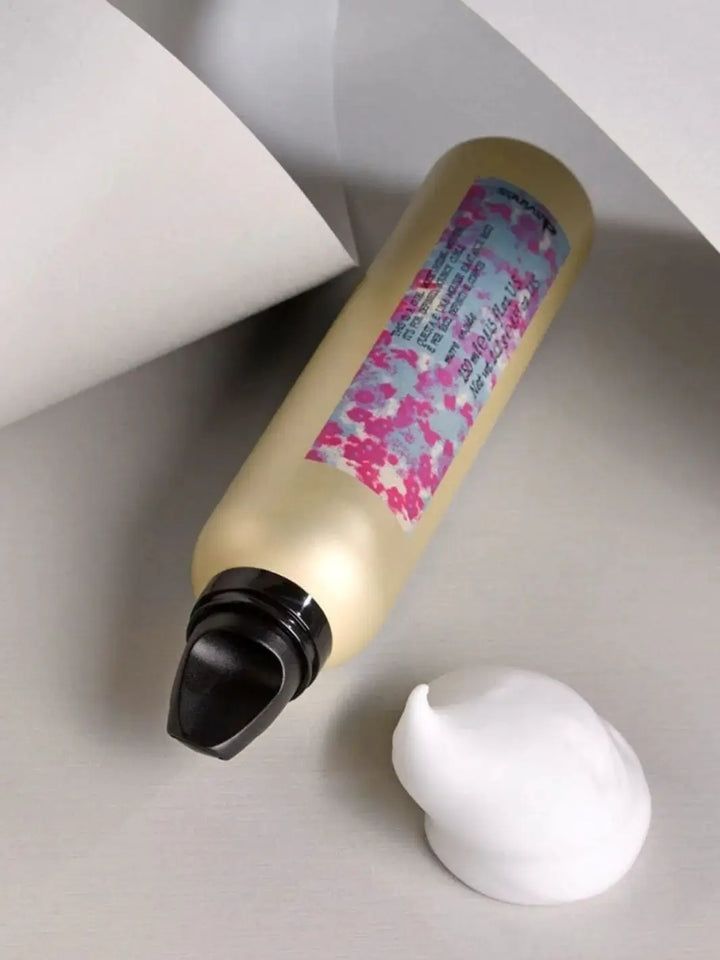 A gold aerosol can of Davines This is a Curl Moisturizing Mousse lies flat next to a pristine mound of white foam on a neutral grey surface | Twentyseven Toronto