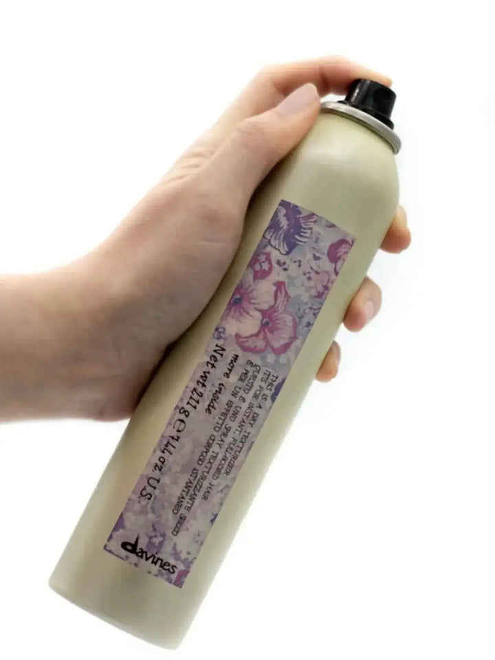 A hand holds a gold aerosol can of Davines This is a Dry Texturizer Spray featuring a purple floral label against a plain white background | Twentyseven Toronto