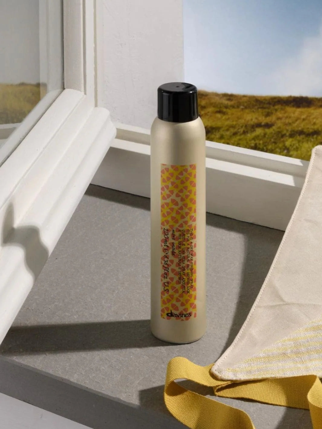 A beige aerosol can of Davines This is a Dry Wax Finishing Spray stands on a grey window sill next to a white window frame with an outdoor landscape in the background | Twentyseven Toronto