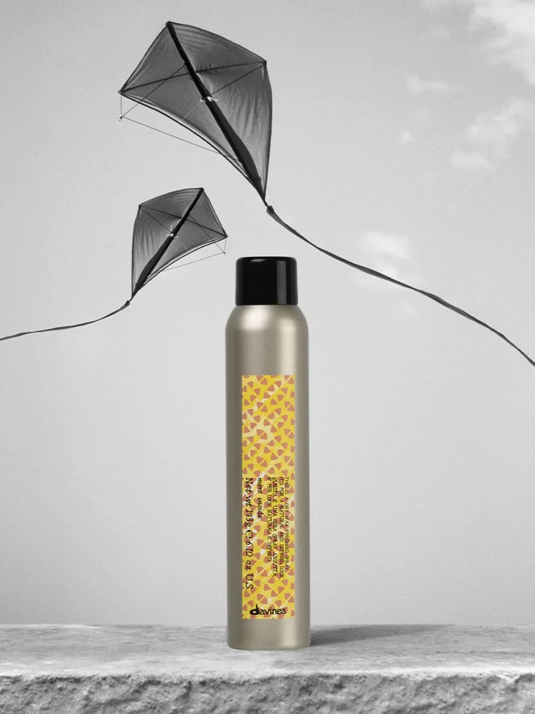 A beige aerosol can of Davines This is a Dry Wax Finishing Spray stands upright on a textured surface beneath two black kites flying in a cloudy sky | Twentyseven Toronto