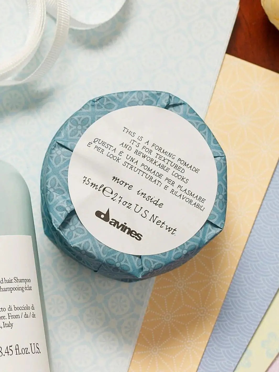 A round container of Davines This is a Forming Pomade, neatly wrapped in blue patterned paper with a white circular label, sits on a flat surface among colorful papers | Twentyseven Torotno