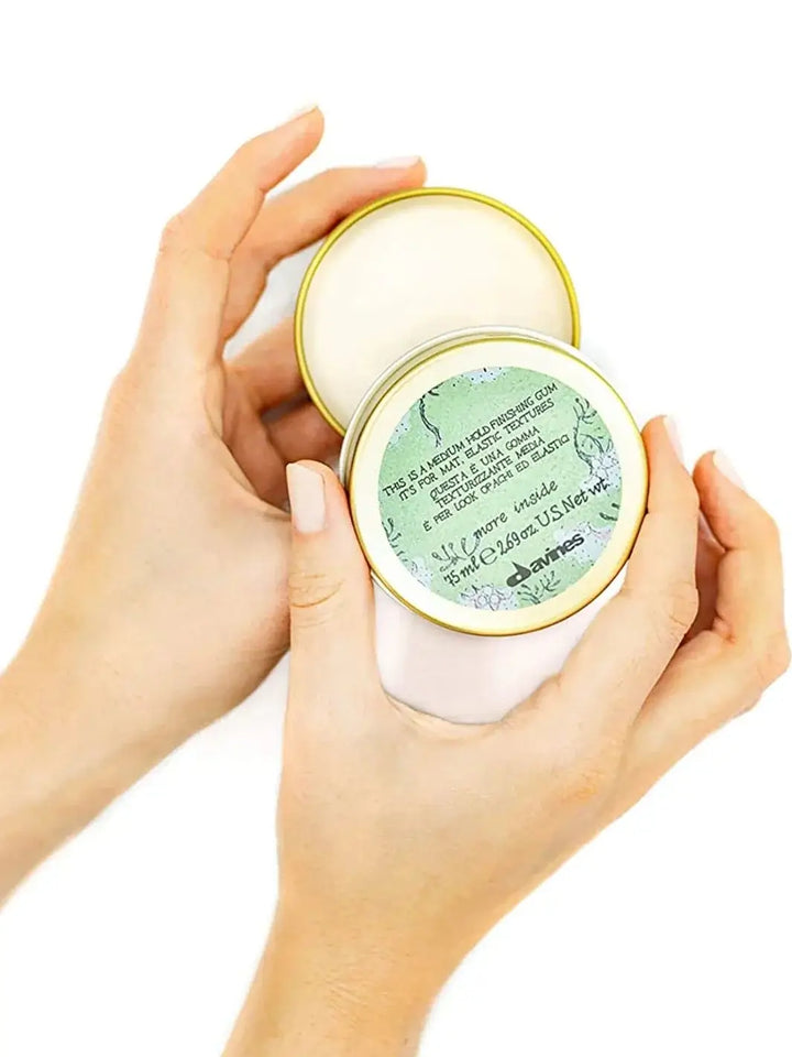 A pair of hands holds an open gold metal tin of Davines This is a Medium Hold Finishing Gum, showing the green patterned product label on the lid against a white background | Twentyseven Toronto