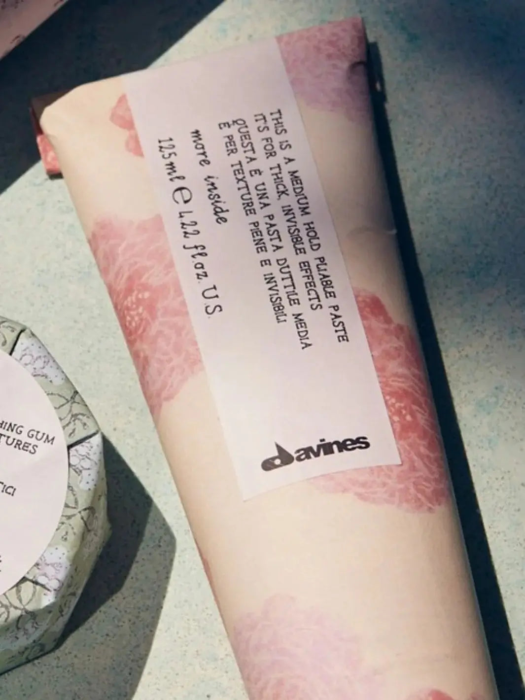 A close-up shot shows the pink floral paper wrapping of a Davines This is a Medium Hold Pliable Paste tube resting on a speckled surface | Twentyseven Toronto