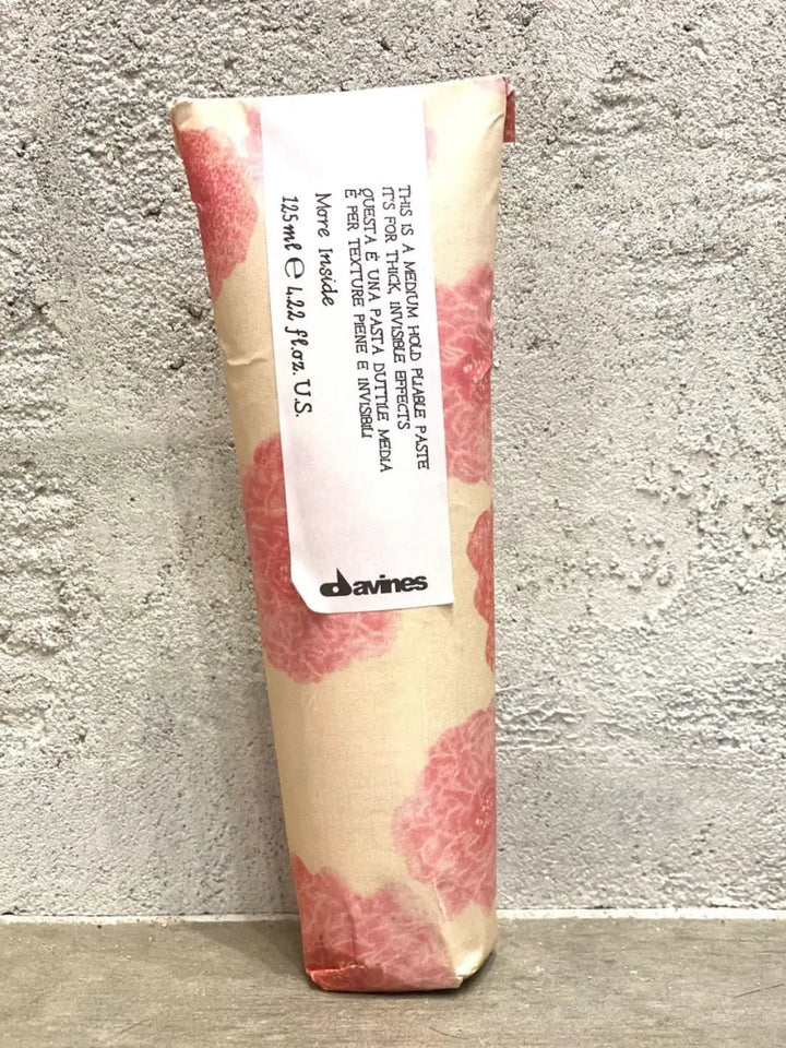 A single tube of Davines This is a Medium Hold Pliable Paste in its floral paper wrapping stands vertically against a textured grey concrete wall | Twentyseven Toronto