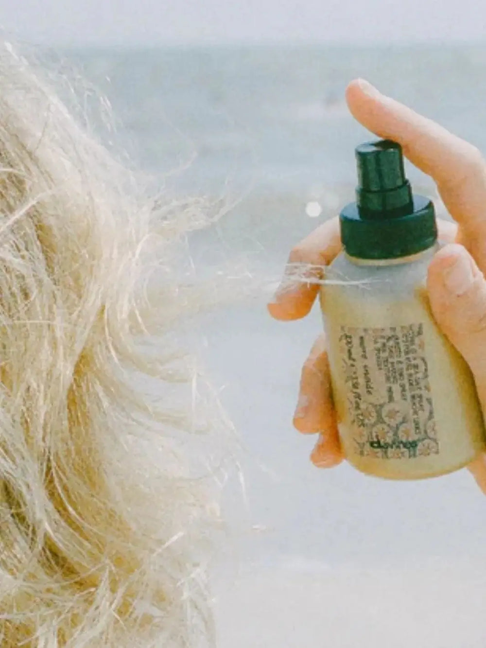 A hand sprays a bottle of Davines This is a Sea Salt Spray onto a section of wavy blonde hair with a blurry beach backdrop | Twentyseven Toronto
