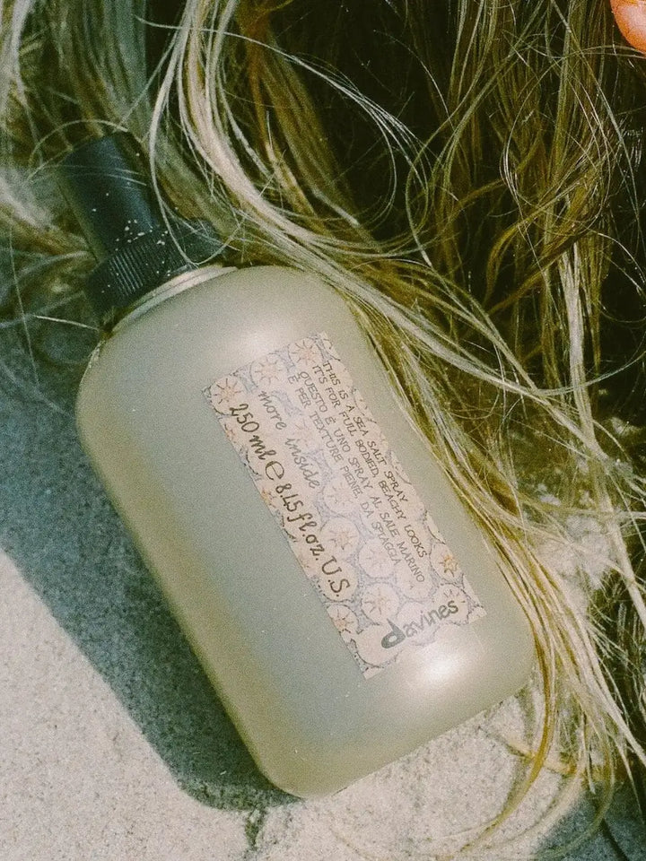 A bottle of Davines This is a Sea Salt Spray rests on the sand next to strands of textured blonde hair | Twentyseven Toronto