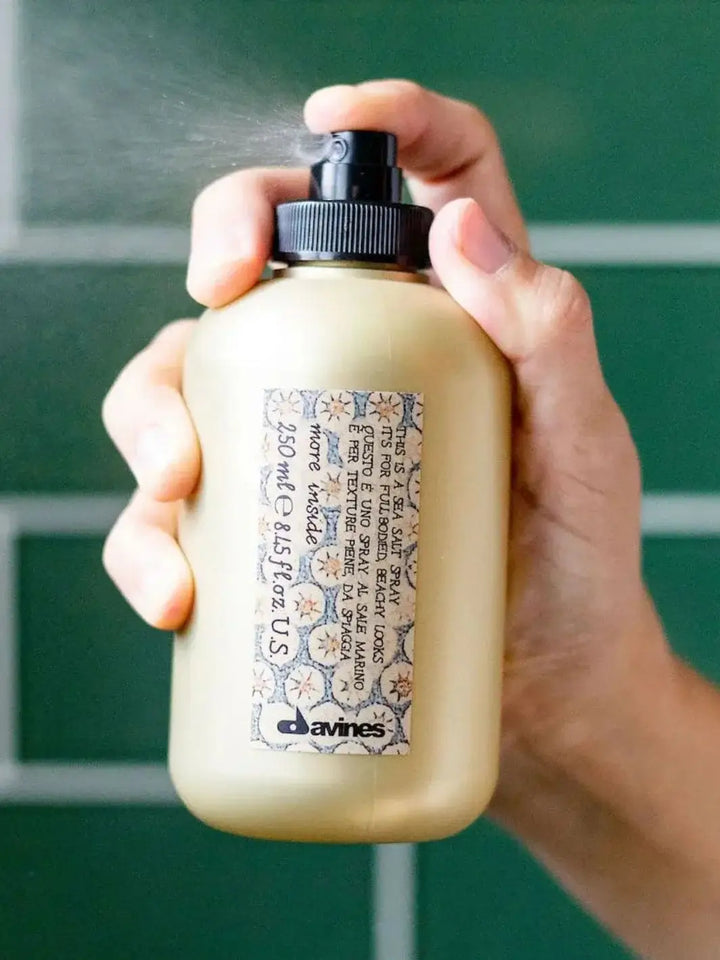 A hand presses the nozzle of a Davines This is a Sea Salt Spray bottle, releasing a fine mist against a green tiled wall | Twentyseven Toronto