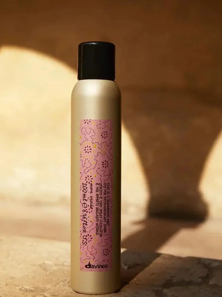 A gold aerosol can of Davines This is a Shimmering Mist with a pink patterned label stands on a textured stone surface, casting a shadow in warm sunlight | Twentyseven Toronto