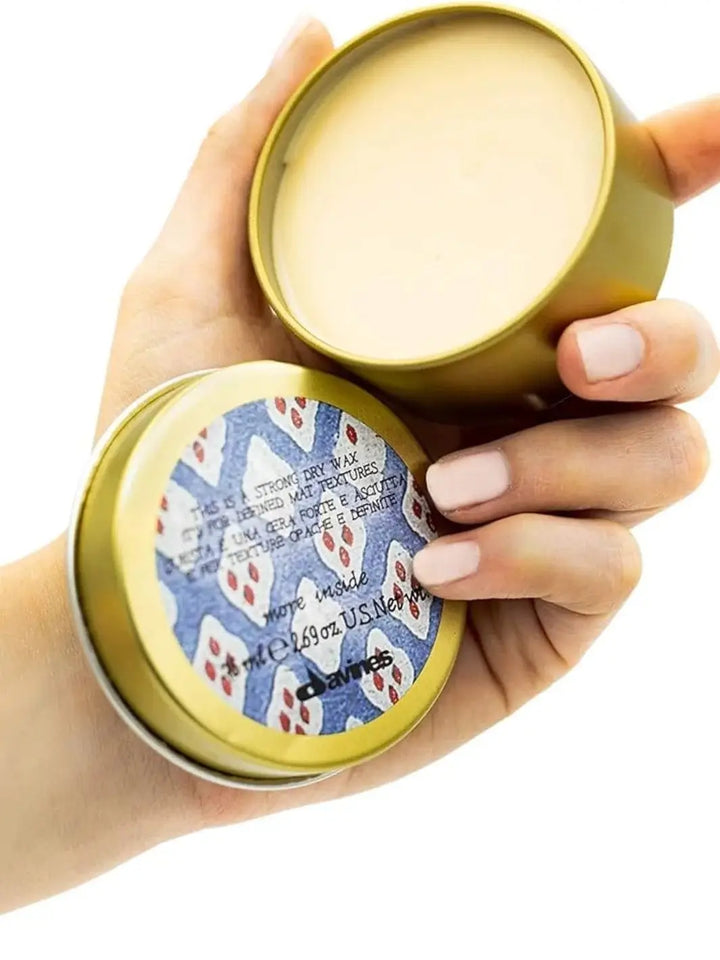 A hand holds an open gold metal tin containing Davines This is Strong Dry Wax, showing the textured product inside and a blue-and-white patterned label on the lid against a white background | Twentyseven Toronto
