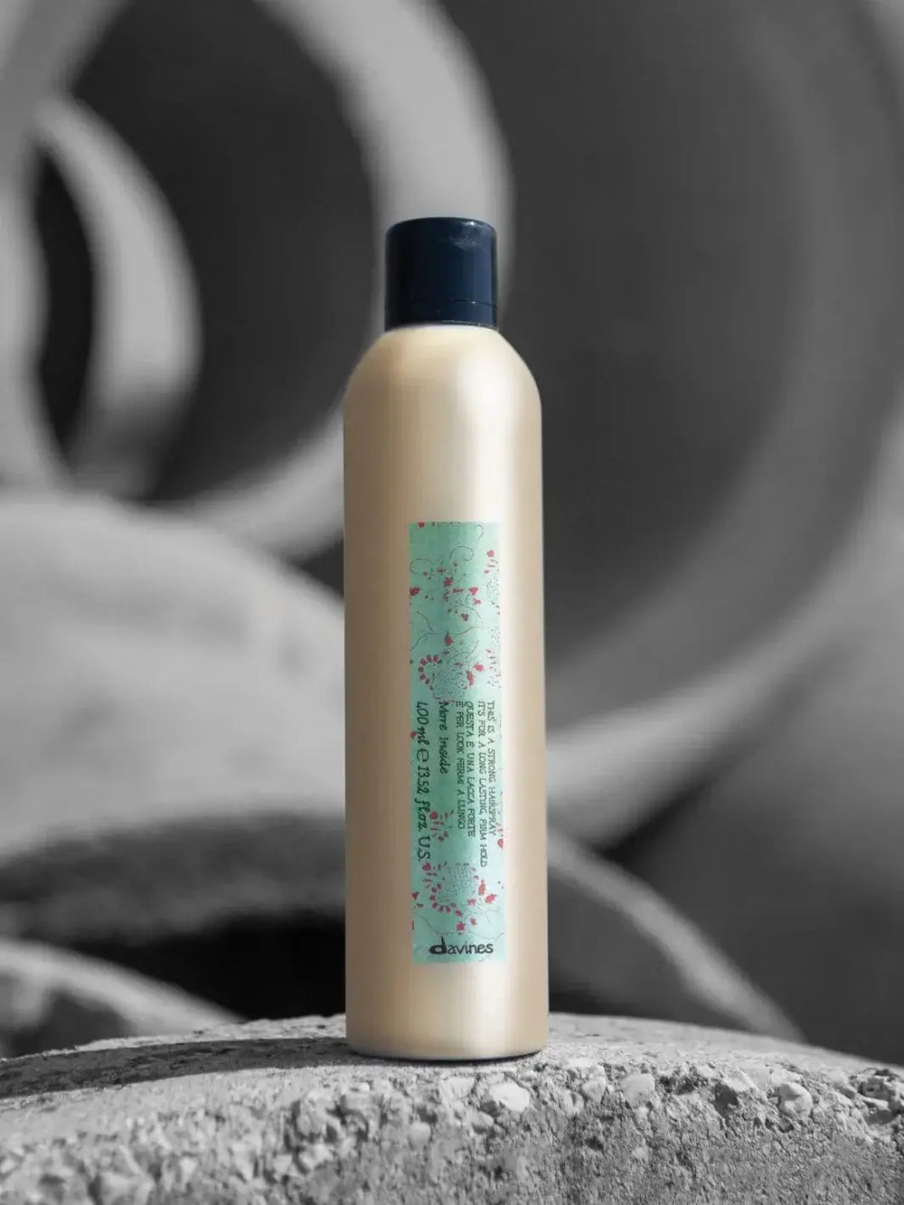 A gold aerosol can of Davines This is a Strong Hold Hairspray featuring a green floral label stands on a textured stone surface with a blurred grey background | Twentyseven Toronto