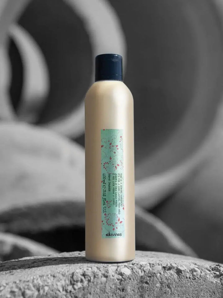 A gold aerosol can of Davines This is a Strong Hold Hairspray featuring a green floral label stands on a textured stone surface with a blurred grey background | Twentyseven Toronto