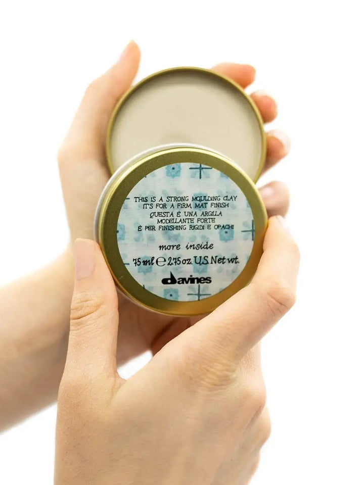 A pair of hands holds an open gold metal tin of Davines This is a Strong Moulding Clay, displaying the blue-patterned product label on the lid against a white background | Twentyseven Toronto
