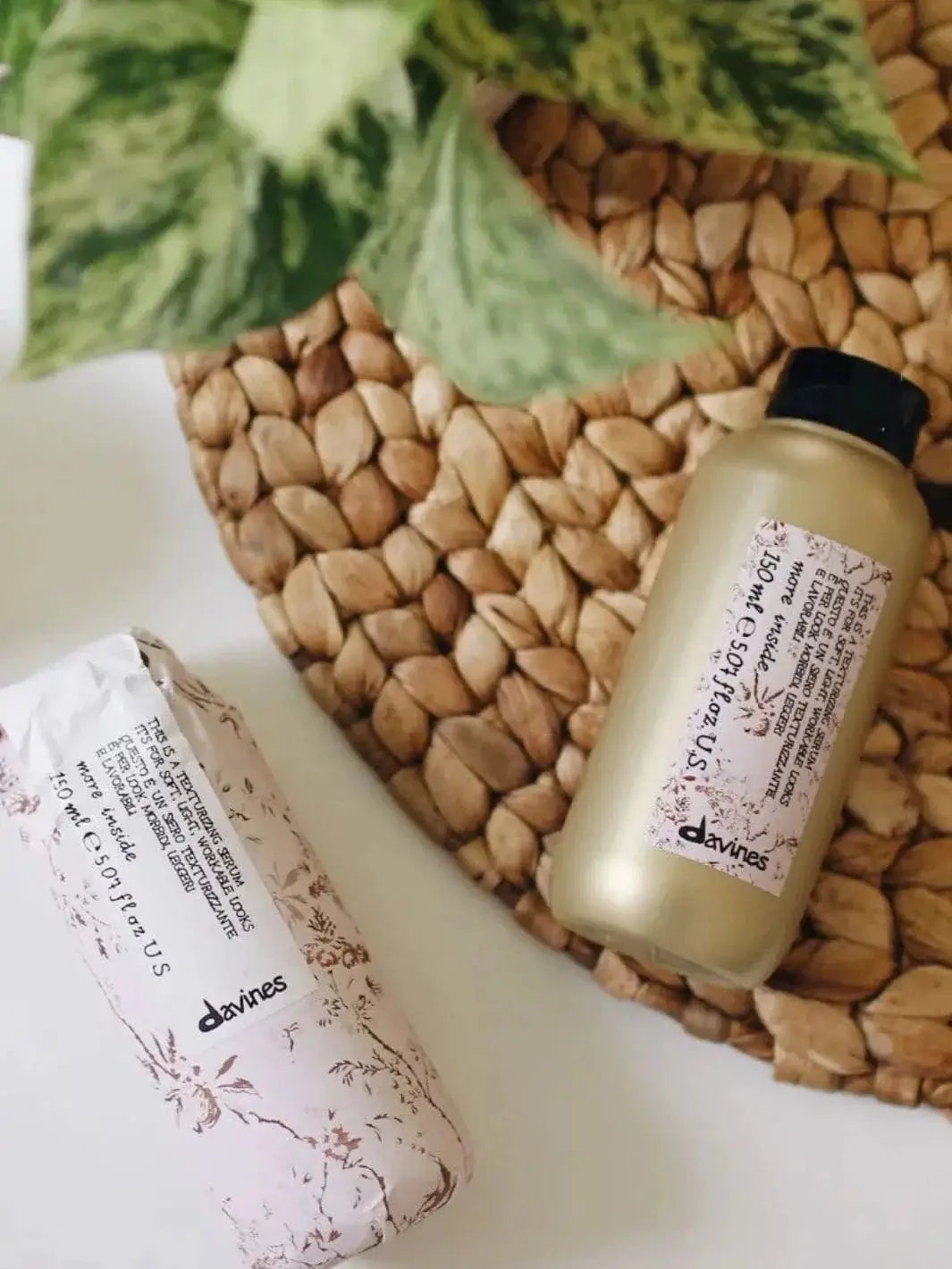 A bottle of Davines This is a Texturizing Serum rests on a woven placemat next to its matching floral paper packaging | Twentyseven Toronto