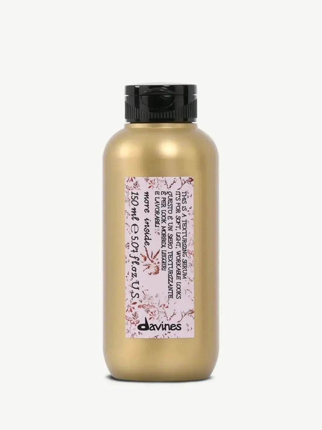 A gold bottle with a black flip-top cap displays the pink floral label of Davines This is a Texturizing Serum against a plain off-white background | Twentyseven Toronto