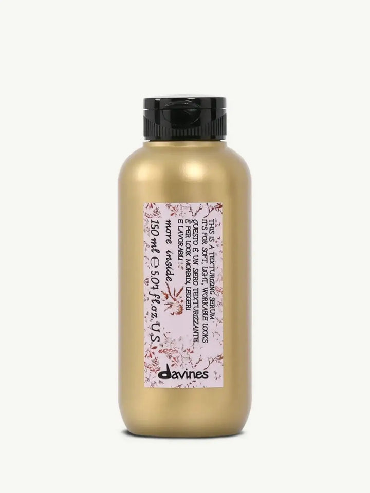 A gold bottle with a black flip-top cap displays the pink floral label of Davines This is a Texturizing Serum against a plain off-white background | Twentyseven Toronto