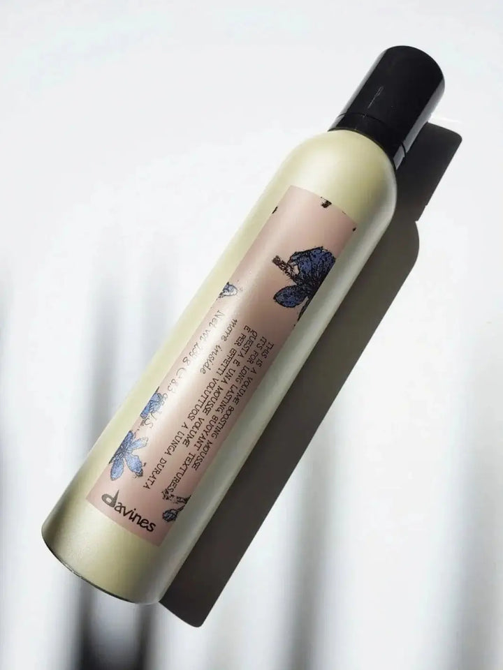 A gold aerosol can of Davines This is Volume Boosting Mousse with a pink and blue floral label lies diagonally on a bright white surface in direct sunlight | Twentyseven Toronto