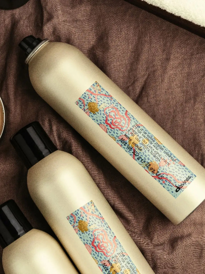 Three gold aerosol cans of Davines This Is a X-Strong Hairspray with blue, pink, and gold patterned labels are arranged diagonally on a textured brown fabric | Twentyseven Toronto