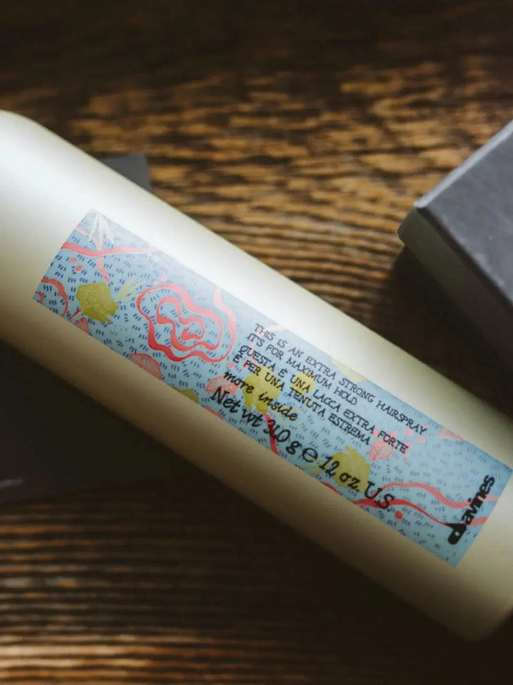 A close-up shot captures a gold aerosol can of Davines This Is a X-Strong Hairspray lying diagonally across a dark wooden surface | Twentyseven Toronto