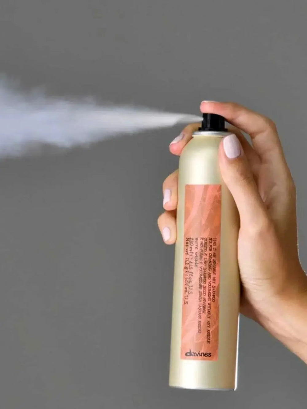 A hand presses the nozzle of a beige canister of Davines This is an Invisible Dry Shampoo, releasing a fine white mist against a solid gray background | Twentyseven Toronto