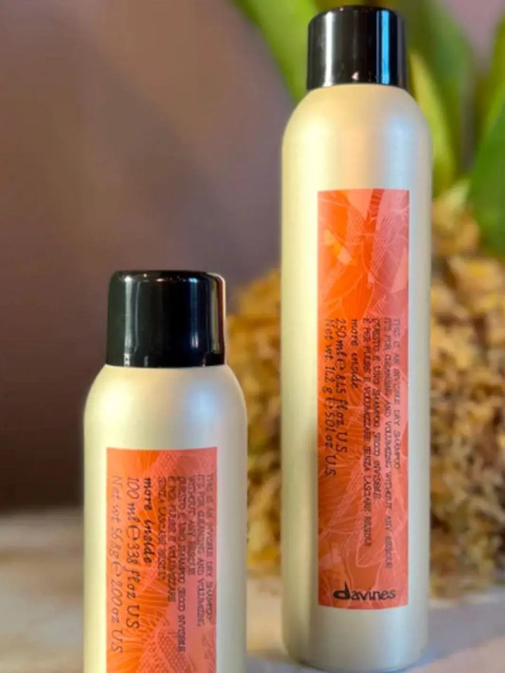 Two different sizes of Davines This is an Invisible Dry Shampoo canisters stand side-by-side with a blurred green and brown plant in the background | Twentyseven Toronto
