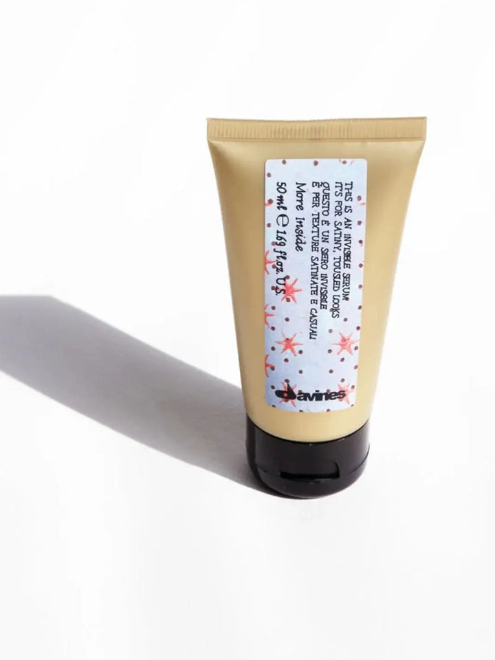 A gold squeeze tube of Davines This is an Invisible Serum featuring a star-patterned label stands on a white surface casting a shadow | Twentyseven Toronto