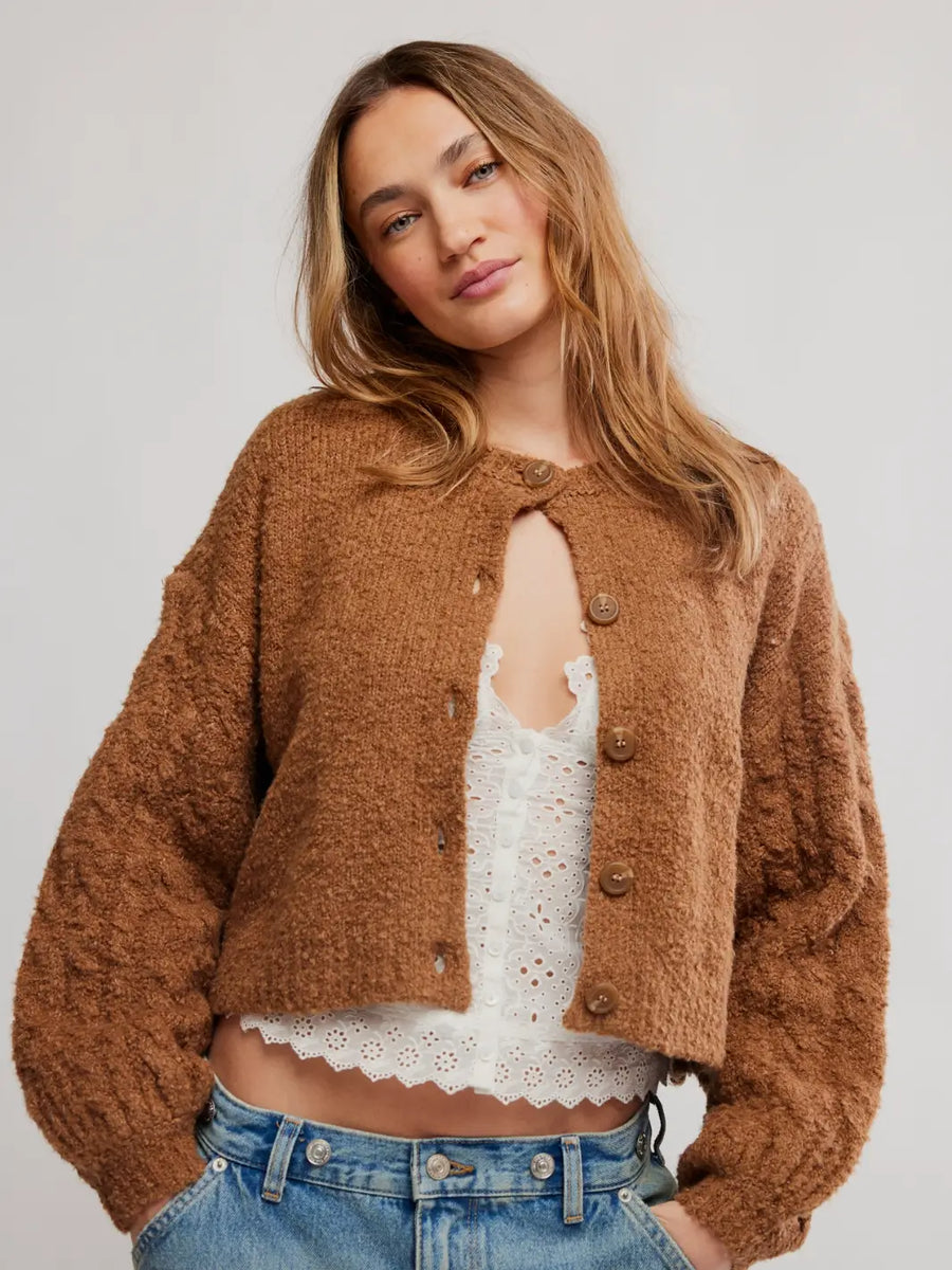 A woman wearing a cropped, fuzzy brown button-front Thistle Cardi in Macadamia from Free People over a white lace-trim camisole and blue jeans, photographed against a plain light gray studio background | Twentyseven Toronto