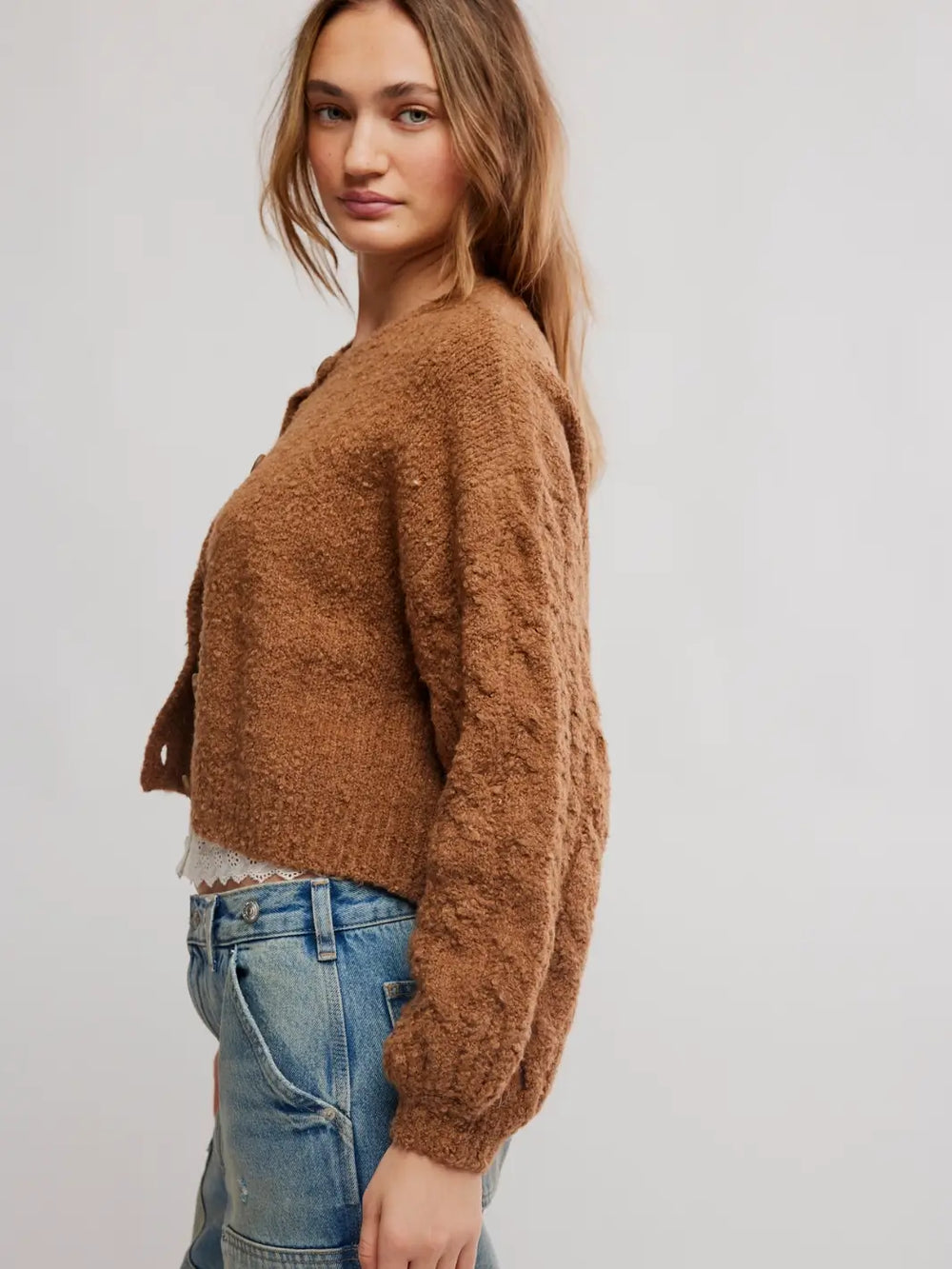 A woman wearing a brown fuzzy cropped Thistle Cardi in Macadamia from Free People and blue jeans, shown from a side profile against a light gray studio background | Twentyseven Toronto
