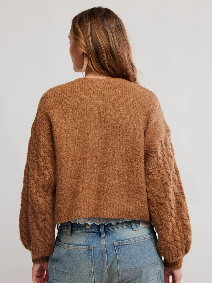 A woman viewed from the back wearing a cropped, textured brown Thistle Cardi in Macadamia from Free People with balloon sleeves and blue jeans, set against a plain light gray studio background | Twentyseven Toronto