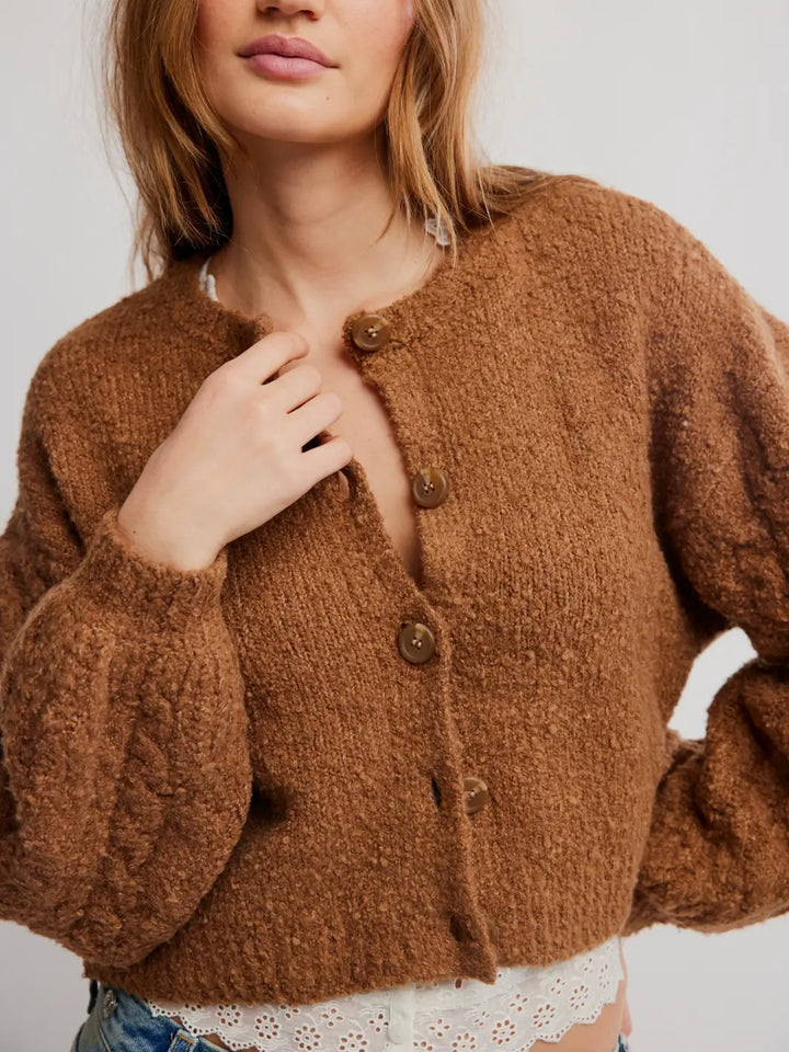 A close-up of a woman wearing a brown fuzzy knit Thistle Cardi in Macadamia from Free People with brown buttons, styled with a white lace top and blue jeans against a light gray background | Twentyseven Toronto