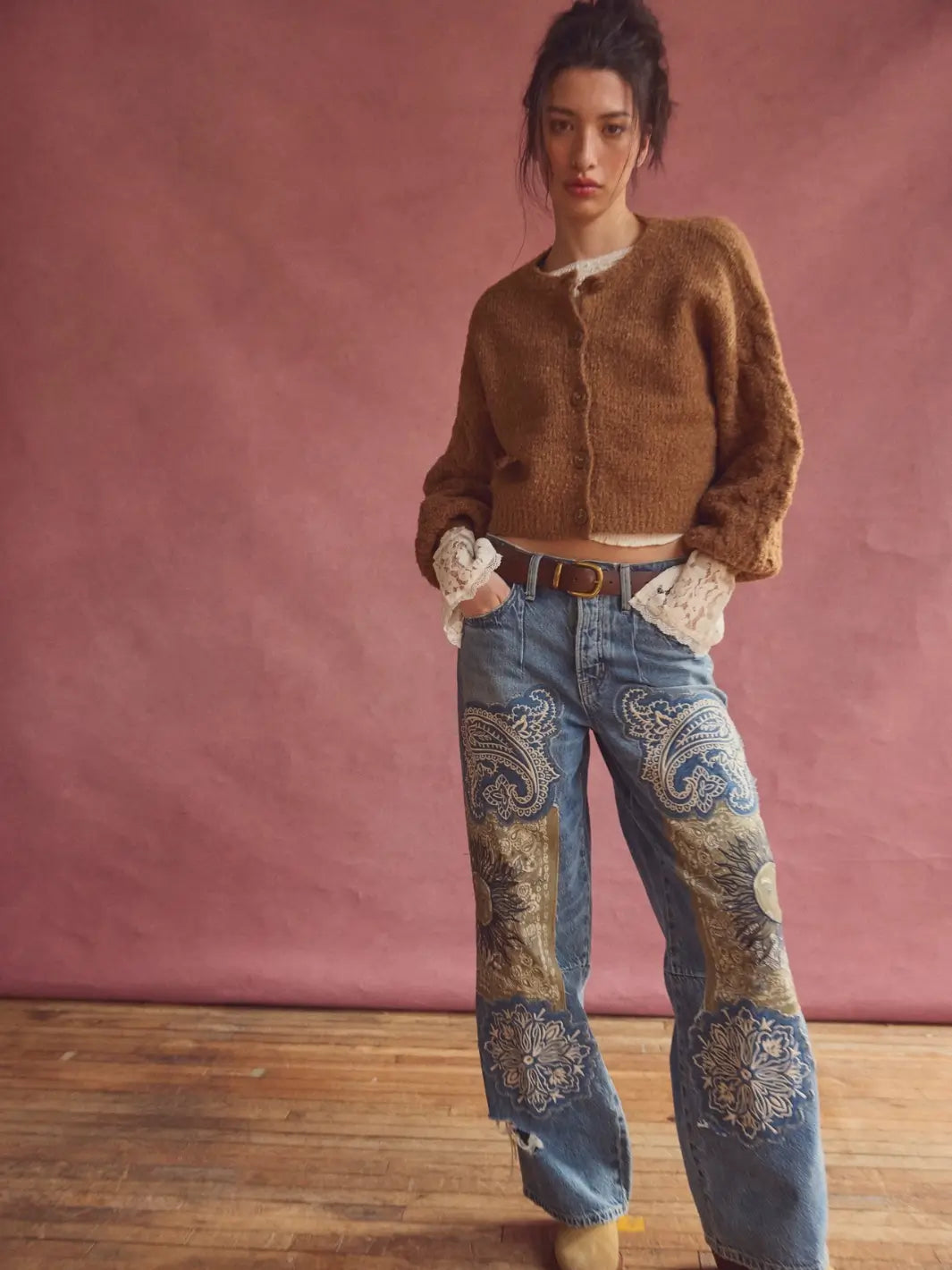 A model stands against a dusty pink studio backdrop wearing a cropped brown fuzzy Thistle Cardi in Macadamia from Free People, wide-leg blue jeans with ornate paisley embroidery on the lower legs, and tan boots | Twentyseven Toronto
