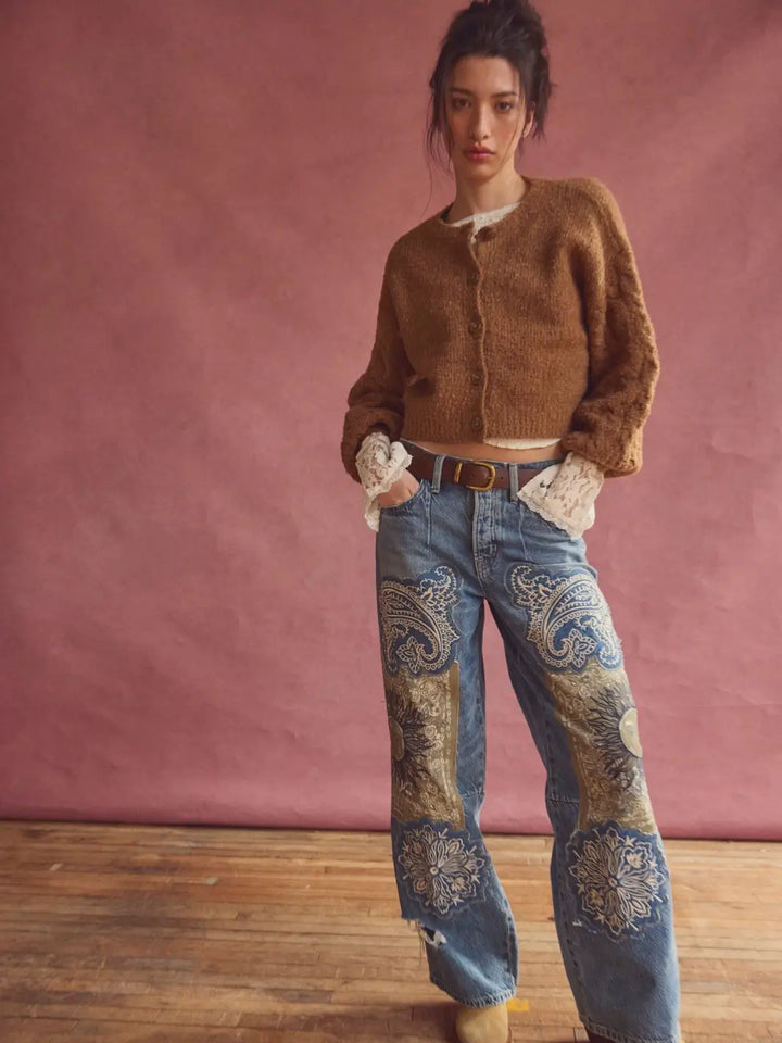 A model stands against a dusty pink studio backdrop wearing a cropped brown fuzzy Thistle Cardi in Macadamia from Free People, wide-leg blue jeans with ornate paisley embroidery on the lower legs, and tan boots | Twentyseven Toronto